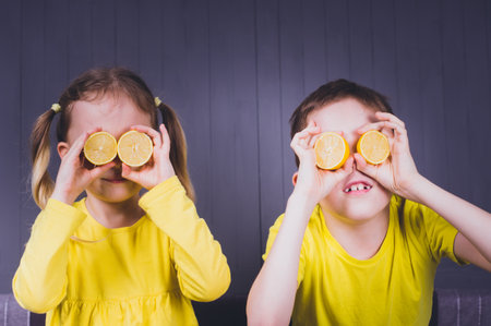 Little smiling cute girl and boy in yellow shirts holding halves of fresh sour lemon fruit near eyes and over gray wooden background.の写真素材