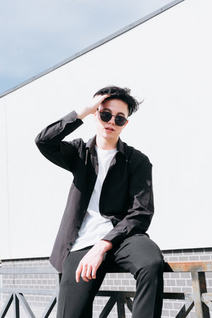 handsome young man in a black shirt standing against the backdrop of city buildings and looking through black sunglassesの写真素材