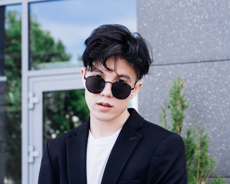 handsome young man in a black shirt standing against the backdrop of city buildings and looking through black sunglassesの写真素材