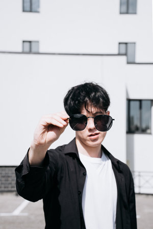 handsome young man in a black shirt standing against the backdrop of city buildings and looking through black sunglassesの写真素材