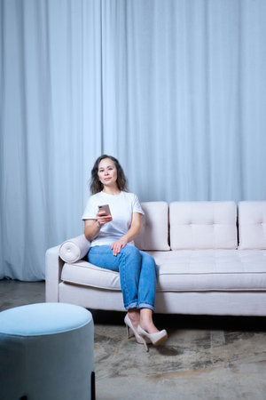a girl in blue jeans and a white t-shirt looks at the phone while sitting on a light-colored sofaの写真素材