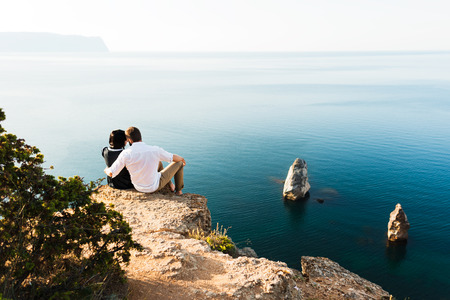 Guy and girl sitting on a cliff by the sea. Honeymoon. Honeymoon trip. Boy and girl at the sea. Man and woman traveling. Couple hugs. Couple kissing. Newly married couple. Loversの写真素材