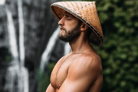 Portrait of a sports guy in an Asian conical hat. Portrait of a man in profile. Portrait of a bearded man. A man meditates in a Balinese conical hat. Beautiful male portrait. A man symbolizing calmの写真素材