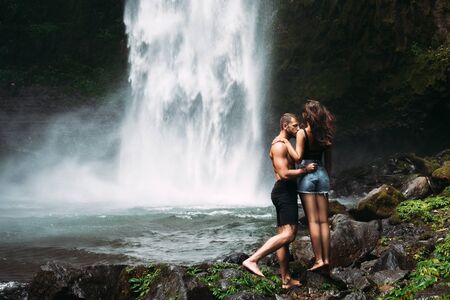 A man of athletic build kisses a beautiful girl at the waterfall. Honeymoon trip. Declaration of...