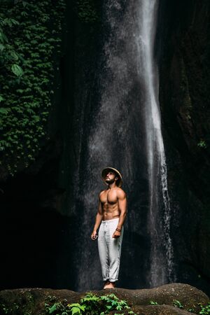 A man of athletic build at the waterfall. A man travels the world. Man at the waterfall. Travel to Bali Indonesia. A man in a conical hat and white pants at the waterfall. Lone traveler.の写真素材