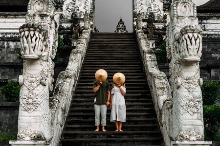 A couple stands on the stairs of the Baltic temple and cover their faces with rice caps. Man and woman traveling in Indonesia. Couple at the Bali gate. The couple travels the world. Tourists in Baliの写真素材