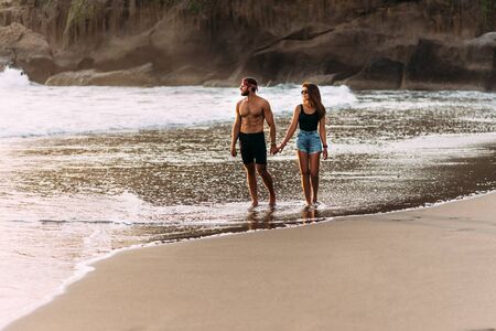 Happy couple strolling along a sandy beach. Couple in love at sunset by the sea. Vacation on the island of Bali. Romantic couple enjoying a beach walk at sunset. Lovers on the coast. Honeymoon tripの写真素材