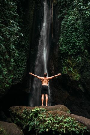 Back view of a man standing by waterfalls with arms outstretched. Man at waterfall raising his hands in feeling closer to nature. Man at the waterfall. Travel to Bali. Man on the background of natureの写真素材