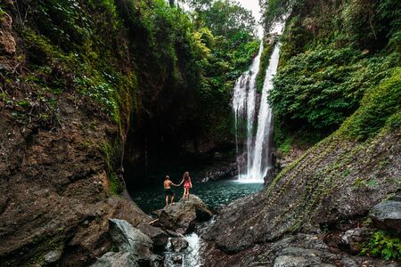 Lovers at the waterfall, rear view. Couple admiring a beautiful waterfall in Indonesia. Couple on vacation in Bali. Honeymoon trip. The couple is traveling in Asia. Vacation on the island of Baliの写真素材