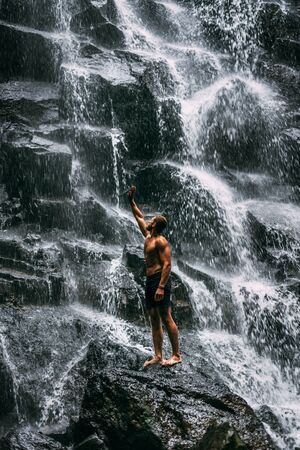 Back view of a man standing by waterfalls with arms outstretched. Man at waterfall raising his hands in feeling closer to nature. Man at the waterfall. Travel to Bali. Man on the background of natureの写真素材