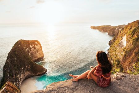 Beautiful girl sitting on a high rock and looking out to sea, rear view. Girl sits on the edge of cliff. The girl of the sea. Seascape. Girl at sunset. Sea tour. Nusa Penida Bali Indonesia. Copy spaceの写真素材