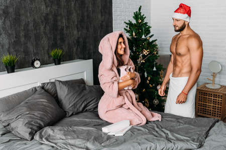 A man makes a new year's gift to his woman. A loving couple enjoys the new year holiday and give each other gifts. A man in a Christmas hat gives a gift to a woman. New year gift. New year at homeの写真素材