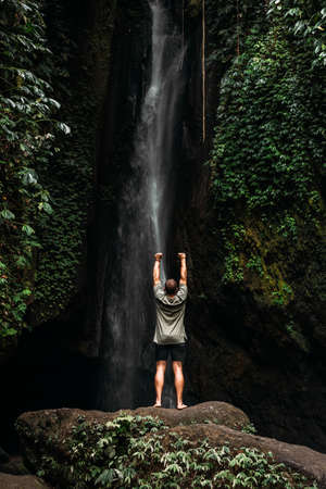 A man stands at the waterfall with his hands raised. Man at waterfall raising his hands in feeling...