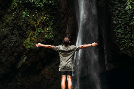 A man stands at the waterfall with his hands raised. Man at waterfall raising his hands in feeling closer to nature. Travel to Bali. Man on the background of nature. Copy space. Vertical photoの写真素材