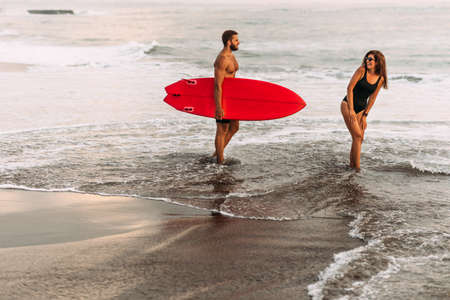 Beautiful couple have fun at the sea. Couple in love on the beach with a surfboard at sunset. Beautiful couple on the island of Bali. Couple of surfers walking on coast in Indonesia. Vacation at seaの写真素材