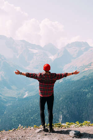 Back view of a man standing against the background of mountains with his arms outstretched. The man in the mountains raises his hands. Man on the background of mountains. Travel to the mountainsの写真素材
