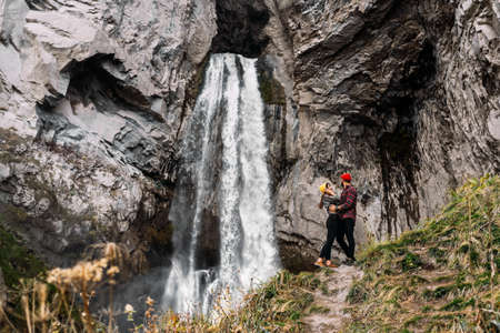 Stylish couple at the waterfall. A man and a woman traveling in the Caucasus. A couple in love is traveling in Russia. Brightly dressed couple of tourists at the foot of a large waterfall. Copy spaceの写真素材