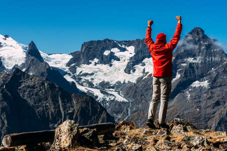 A man in a red jacket on top of a mountain, rear view. A tourist stands on top of a mountain and enjoys what is happening. Active sport concept. The man reached the top of the mountain. Copy spaceの写真素材