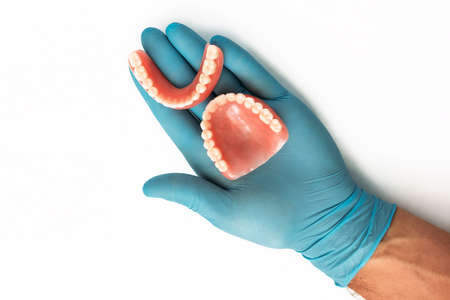 The doctor's hand in a blue glove holds a denture. Dental prosthesis in the hands of the doctor close-up. Dentist holding ceramic dental bridge. Dentistry conceptual photo. Prosthetic dentistry.の写真素材