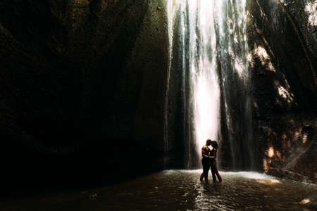 Beautiful couple in a cave with a waterfall. Athletic man and woman under the streams of a waterfall. A sexy couple under a tropical waterfall in twilight light. Tukad Chepung Waterfall. Copy spaceの写真素材