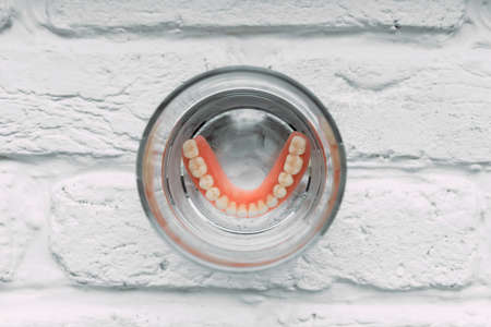 A denture in a glass of water. Dental prosthesis care. Full removable plastic denture of the jaws. Acrylic denture. Lower jaw with false teeth. Dentures or false teeth, close-up.の写真素材