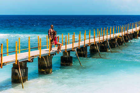 A beautiful couple on vacation strolling along a wooden pier above the tropical, turquoise ocean in Nusa Penida, Indonesia. Couple on vacation. Lovers on the coast. Lovers walk along the pierの写真素材