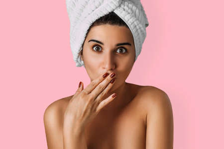 Portrait of a young attractive woman with a towel on her head on an isolated background. A girl with a towel on her head stands on a pink background, covering her mouth with her hand, close-up.の写真素材
