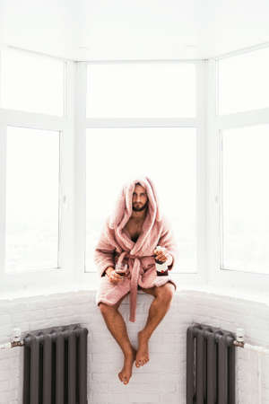 A man in a dressing gown is sitting on the windowsill. A man in a rabbit costume. A man in a pink robe. A man with a bottle of alcohol at the window. Vertical photoの写真素材