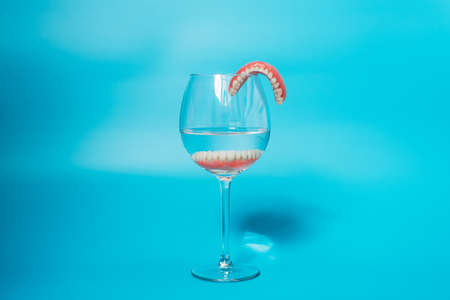 Dental prosthesis care. Full removable plastic denture of the jaws. A set of dentures in a glass of water on a blue background. Two acrylic dentures. Dentures or false teeth, close-up. Copy spaceの写真素材