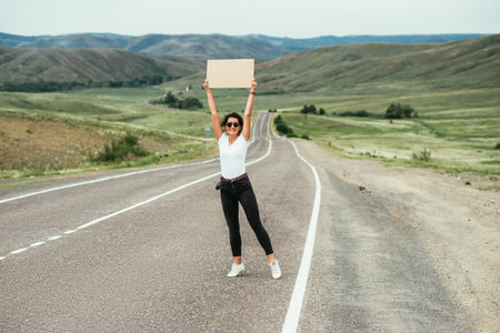 The girl is hitchhiking. Passion for travel, the concept of auto travel. A hitchhiking trip. A woman tries to stop a car on the road with an empty cardboard sign, a mock-up. Traveling by car.の写真素材