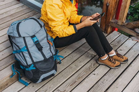 A traveler with a phone, close-up. A tourist girl in a yellow jacket with a backpack holds a phone in her hands. Hiking woman using smart phone, travel and active lifestyle concept. Copy spaceの写真素材