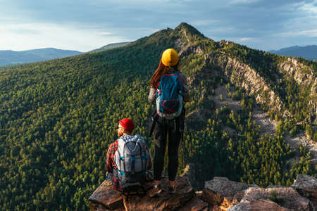 A man and a woman with backpacks on the mountain admire the panoramic view. A couple in love on a rock admires the beautiful views. Travelers in the mountains at sunset. A couple on a hike. Copy spaceの写真素材