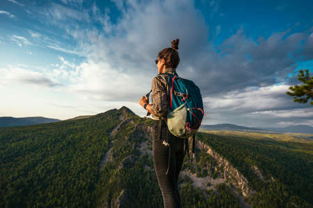 A young tourist girl with a backpack enjoys the sunset from the top of the mountain. A traveler on the background of mountains. A tourist with a backpack is standing on the top of the mountainの写真素材
