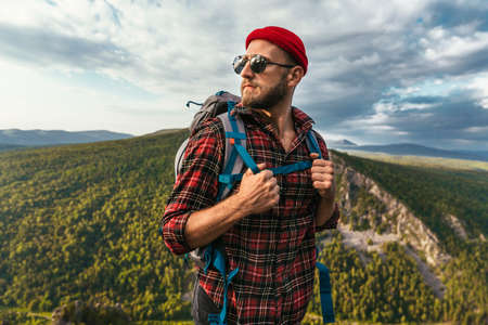 A bearded traveler with a backpack on the top of a mountain. Portrait of a traveler in a red cap and sunglasses. A tourist with a backpack stands against the background of a mountain. Copy spaceの写真素材