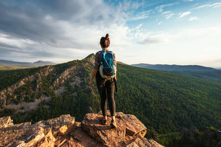 A young tourist girl with a backpack enjoys the sunset from the top of the mountain. A traveler on the background of mountains. A tourist with a backpack is standing on the top of the mountainの写真素材