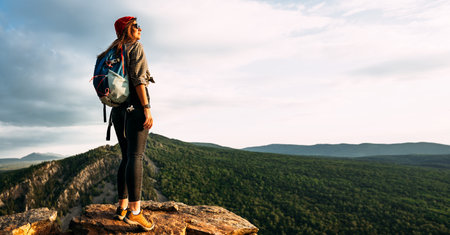 A young female tourist with a backpack admires the sunset from the top of the mountain, panorama. A traveler on the background of mountains. Tourist with a backpack on the background of mountainsの写真素材