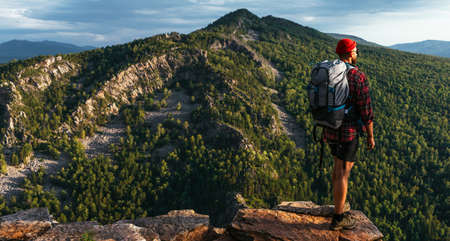 A male tourist with a backpack admires the sunset from the top of the mountain, panorama. A traveler on the background of mountains. Tourist with a backpack on a mountain background. Copy spaceの写真素材