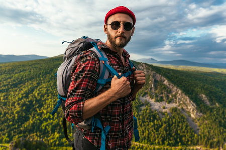 A bearded traveler with a backpack on the top of a mountain. Portrait of a traveler in a red cap and sunglasses. A tourist with a backpack stands against the background of a mountain. Copy spaceの写真素材