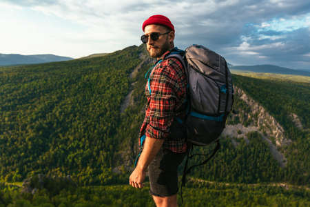 A bearded traveler with a backpack on the top of a mountain. Portrait of a traveler in a red cap and sunglasses. A tourist with a backpack stands against the background of a mountain. Copy spaceの写真素材