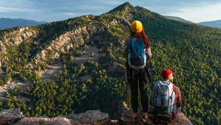 Two travelers on the background of mountains, panorama. Travelers at sunset in the mountains. Tourists with backpacks on the edge of the cliff. A couple admires the sunset from a cliff. Copy spaceの写真素材