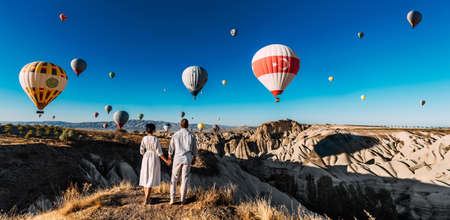A couple in love stands against the background of balloons in Cappadocia, panorama. Travel to the tourist places of Turkey. A man and a woman on a hill look at a large number of flying balloonsの写真素材