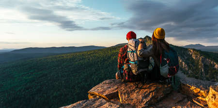 A happy couple in the mountains admires the beautiful views. A man and a woman with backpacks on the mountain admire the panoramic view. Travelers enjoy climbing the mountain at sunset. Copy spaceの写真素材