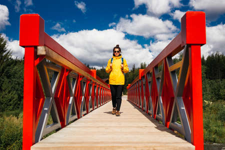 A young tourist girl in a yellow jacket with a backpack is walking along the red bridge. A tourist walks through the national park. A tourist passes the river through the bridge. Copy spaceの写真素材