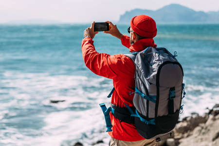 A traveler takes pictures of nature on a smartphone. A traveler with a backpack travels through picturesque places. Tourist by the sea. A traveler on the seashore. A man travels around the islandの写真素材