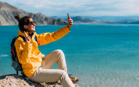 A tourist girl is talking on a video call against the background of the sea. A traveler takes a selfie on her smartphone against the background of the sea. A traveler on the seashore. Copy spaceの写真素材