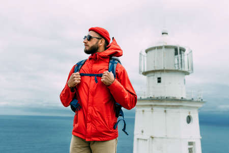 A traveler on the background of a lighthouse by the sea. Portrait of a traveler in red clothes on the coast. A traveler by the ocean. Portrait of a bearded man traveling with a backpack. Lighthouseの写真素材