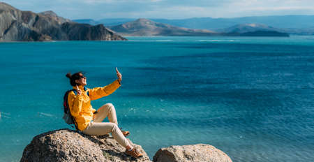 A female traveler is talking on a video call against the background of the sea, panorama. A traveler takes a selfie on her smartphone against the background of the sea. A traveler on the seashore.の写真素材