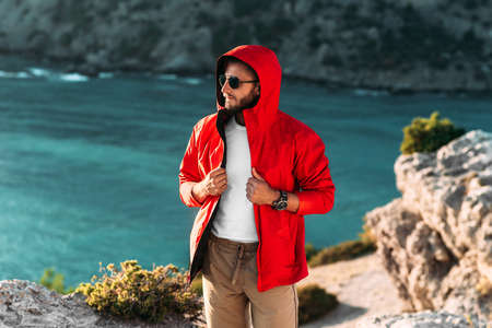 Portrait of a brutal bearded man in a red jacket by the sea. Male portrait on the background of a seascape. A traveler on the background of the sea coast. A man in a jacket at sunsetの写真素材