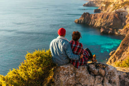 A man and a woman are sitting on the edge of a cliff by the sea. A couple in love at sunset by the sea, rear view. A man and a woman are traveling. A stylish couple meets the sunset on the seashore.の写真素材