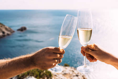Hands holding champagne glasses over the sea. Romantic vacation. Two hands holding champagne glasses on the background of the sea. Toast with champagne glasses on the seashore. Copy spaceの写真素材